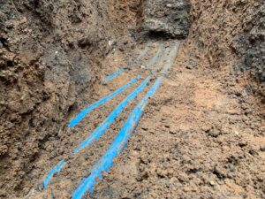 Polybutylene pipe replacement showing new blue PEX or polyethylene water service lines laid in a dirt trench next to the old, buried piping.